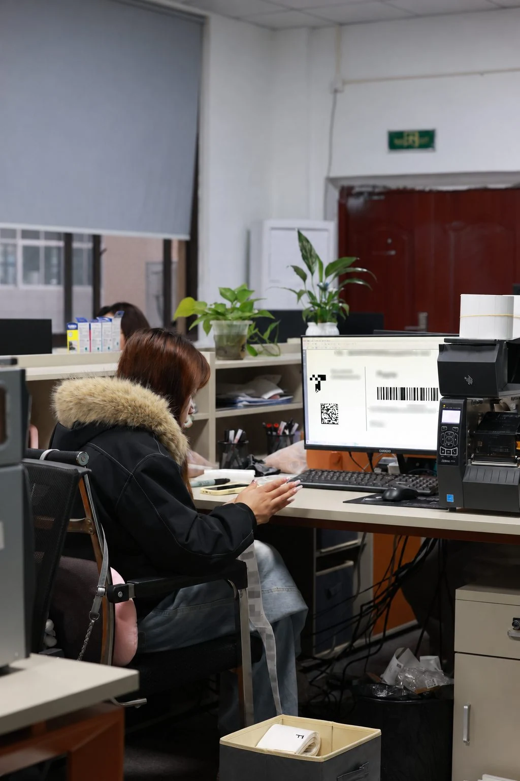 Moving Bags Manufacturing Tiiocti Office Worker Beside A Label Printer Viewing A Label Layout With Visible Barcode Graphics And Selectively Blurred Commercial Text
