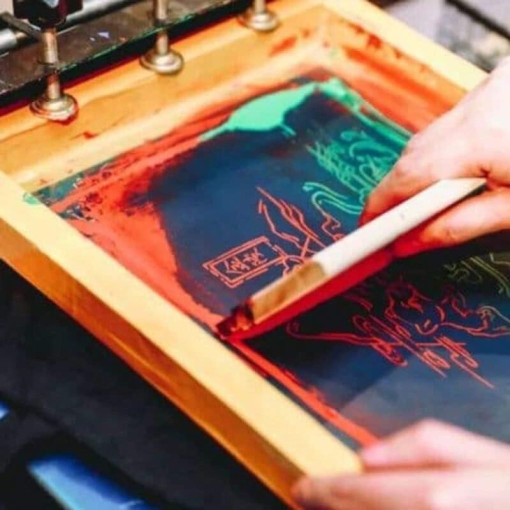 Hands applying ink on a screen printing frame for personalized logo designs on eco-friendly canvas bags.
